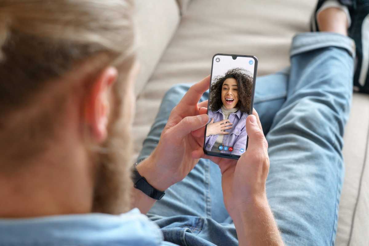 Young man sitting on sofa holding smartphone communicating with african girl friend on mobile screen, making video call using cell phone mobile social media dating app. Video call concept. Over shoulder closeup view